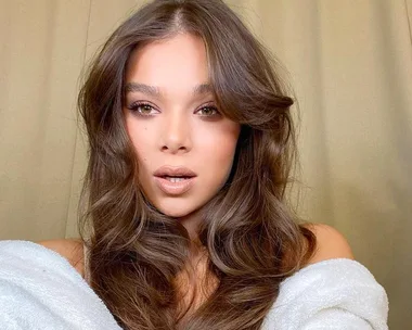 Woman with long, wavy brown hair and neutral makeup, wearing an off-shoulder white garment, in front of a beige background.