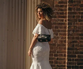 Woman in a white lace dress with an off-shoulder design, standing near a brick wall, holding a black designer handbag.