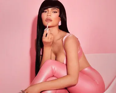 A woman in pink clothing applies lip gloss against a pink background.