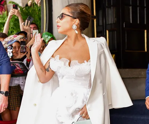 Woman in white dress and jacket, wearing sunglasses and waving to fans outside a building.