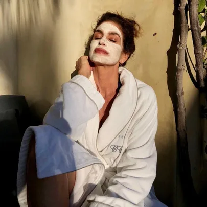 Person in a white robe and facial mask enjoying sunlight, with closed eyes, leaning against a wall.