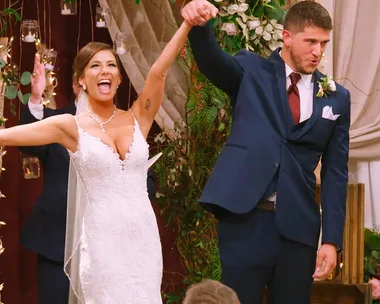 A bride and groom celebrate joyfully holding hands at their wedding, surrounded by flowers and string lights.