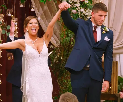 A bride and groom celebrate joyfully holding hands at their wedding, surrounded by flowers and string lights.