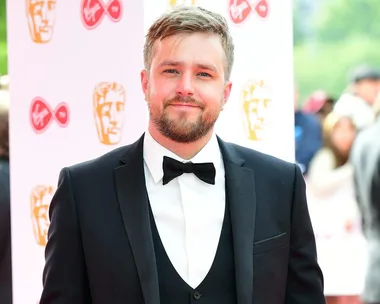 Man in a suit with a bow tie at a red carpet event, smiling and standing in front of a backdrop with logos.