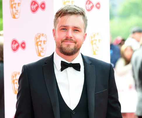 Man in a suit with a bow tie at a red carpet event, smiling and standing in front of a backdrop with logos.