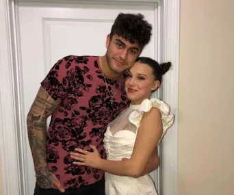 A man in a floral shirt poses with a woman in a white dress, both smiling against a door backdrop.