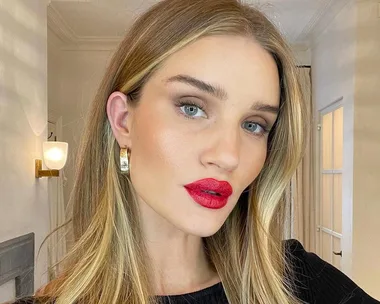 A woman with long blonde hair wears bright red lipstick and hoop earrings, posing indoors with soft lighting.