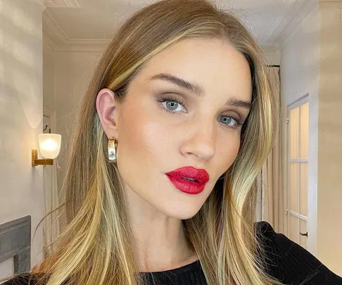 A woman with long blonde hair wears bright red lipstick and hoop earrings, posing indoors with soft lighting.