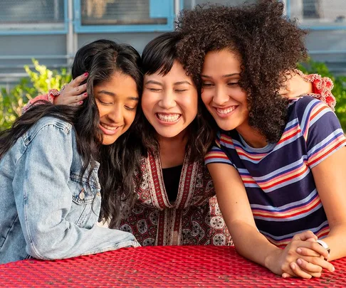 Three young women sit at a red table, smiling and hugging, outdoors in a scene from "Never Have I Ever" Season 2.