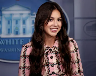 Olivia Rodrigo smiles in a pink plaid jacket during a White House visit, standing in front of a blue background.