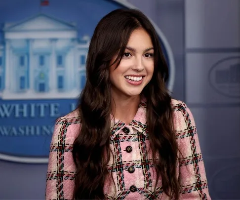 Olivia Rodrigo smiles in a pink plaid jacket during a White House visit, standing in front of a blue background.