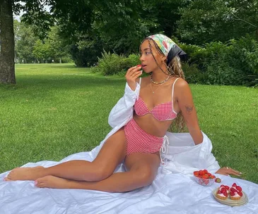 Woman in red checkered bikini and headscarf enjoying strawberries on a picnic blanket in a lush green park.