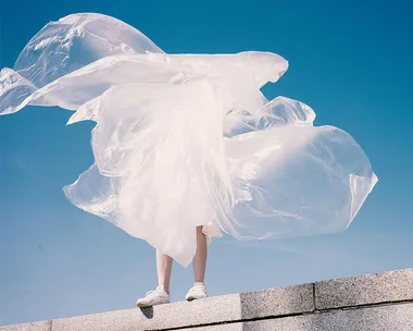 Person standing on a ledge with plastic sheet billowing in the wind against a blue sky.