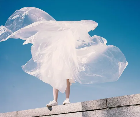 Person standing on a ledge with plastic sheet billowing in the wind against a blue sky.