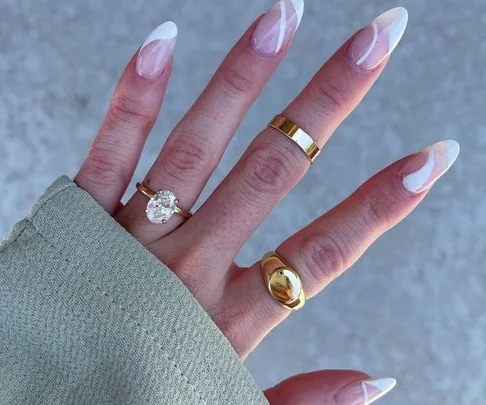 A hand with a gold engagement ring featuring a large oval diamond, wearing a green sleeve and two gold bands on manicured nails.