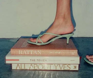 A foot in a green strappy sandal steps on a stack of books, including "Rattan," "The Touch," and "All About Yves."