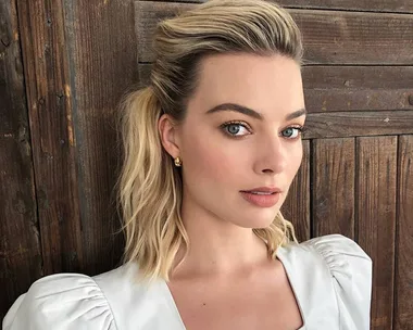 Blonde woman with shoulder-length hair styled in a voluminous, wavy look, wearing a white blouse and gold earrings.