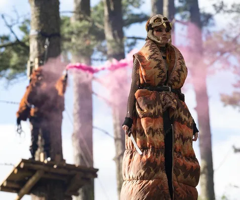 A character in a camo-patterned vest with face paint stands in a forest, surrounded by pink smoke in "Sweet Tooth" Season 2.