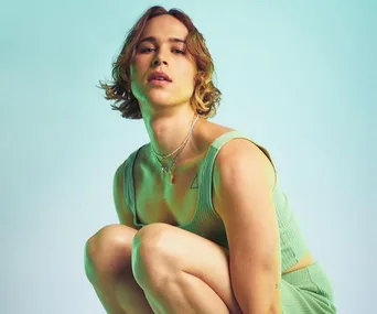 Person with short hair wearing a green outfit, crouching against a teal background.