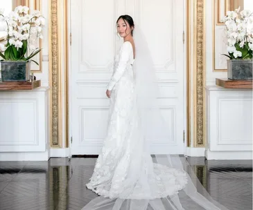 Bride in elegant long-sleeved lace gown and veil, standing indoors with ornate decor and orchids.