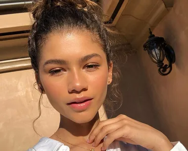 Young woman with hair in a bun, glowing skin, and natural makeup, posing outdoors in sunlight.