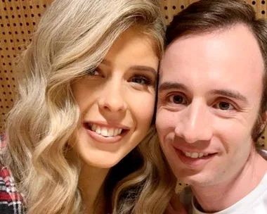 A woman with wavy blonde hair and a man with dark hair smiling closely together against a wooden backdrop.