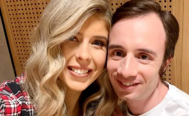 A woman with wavy blonde hair and a man with dark hair smiling closely together against a wooden backdrop.