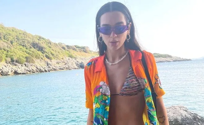 A woman in a colorful shirt and sunglasses stands by a scenic rocky coastline and blue sea.