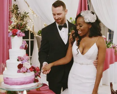 Bride and groom smiling as they cut a wedding cake adorned with pink and red roses, under white and pink drapery.