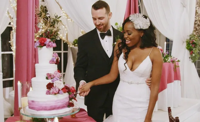 Bride and groom smiling as they cut a wedding cake adorned with pink and red roses, under white and pink drapery.