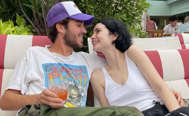 A couple sits close on a striped couch, smiling and looking into each other's eyes, with plants in the background.