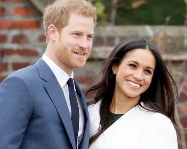 A smiling couple poses outdoors, the man in a blue suit and the woman in a white coat.