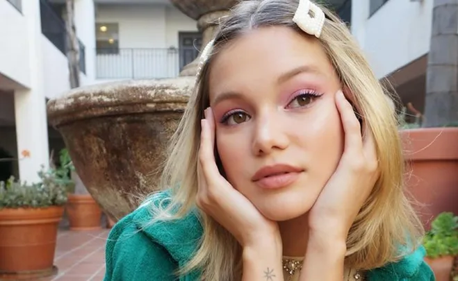 A young woman with blonde hair and pearl hairpin poses outdoors with her hands on cheeks, wearing pink eyeshadow.