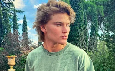 A person with short, tousled hair in a green shirt stands outdoors with trees in the background.