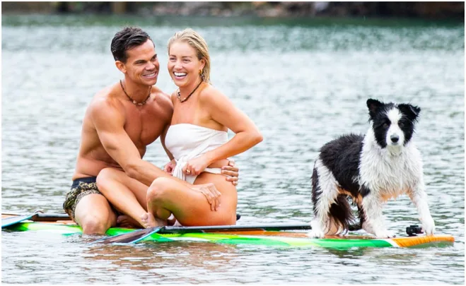 A couple sits happily on a paddleboard in the water, with a dog standing beside them.