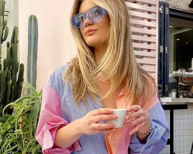 Woman in a color-block shirt holding a coffee cup, sitting outside a café with cacti in the background.