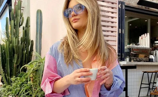 Woman in a color-block shirt holding a coffee cup, sitting outside a café with cacti in the background.