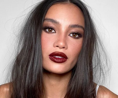 A person with long dark hair, wearing bold eyeliner and deep red lipstick, looking directly at the camera.