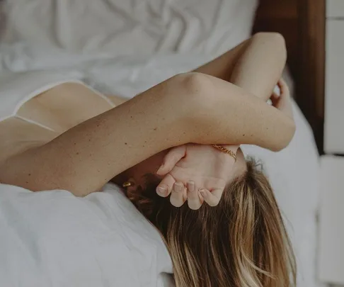 Person lying on a bed, covering their face with arms, wearing a white top, depicting a sense of distress or contemplation.