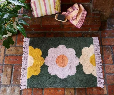 Colorful flower-patterned bath mat on brick floor, with plants and towels nearby.