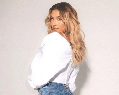 Woman with long, wavy blonde hair wearing a white top and denim jeans, turning slightly to the side.