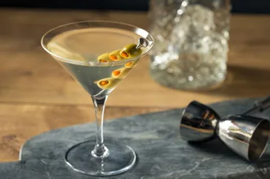 A dirty martini with olives on a skewer in a glass on a slate board, with a shaker in the background.