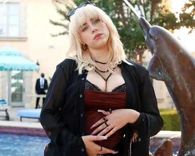 Young woman with blonde shag haircut poses outdoors near a fountain, wearing a black top and jacket.