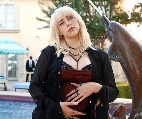 Young woman with blonde shag haircut poses outdoors near a fountain, wearing a black top and jacket.