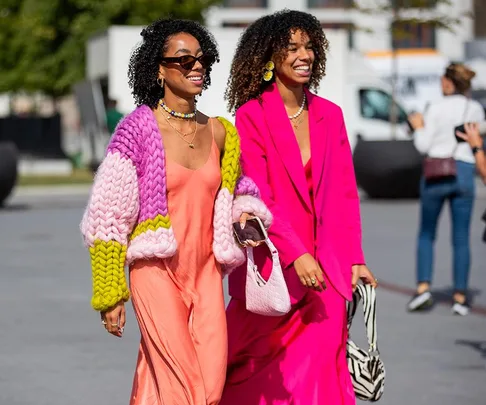 Two women in vibrant outfits, one in a colorful knit cardigan, the other in a bright pink suit, smiling outdoors.