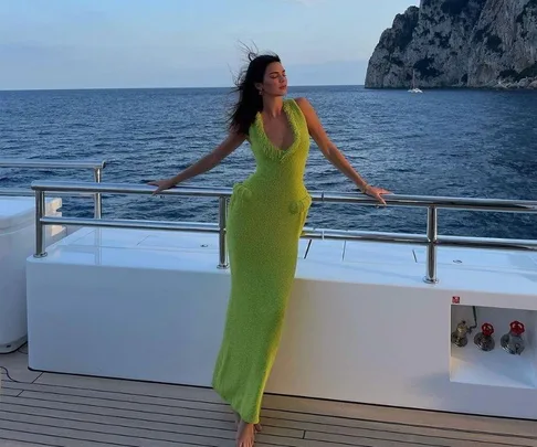 Woman in a spring-green dress poses on a yacht with a scenic ocean and rocky coastline in the background.