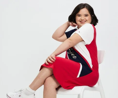 Woman in red, white, and navy dress sitting on a white chair, smiling, with short dark hair and white sneakers.