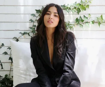 Woman in black blazer seated in front of a white brick wall with green ivy.