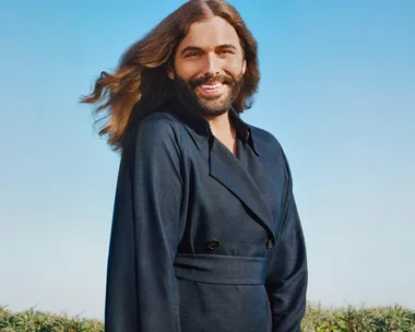 Person with long flowing hair and beard, smiling in an outdoor setting with a blue sky background.