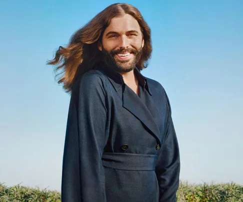 Person with long flowing hair and beard, smiling in an outdoor setting with a blue sky background.